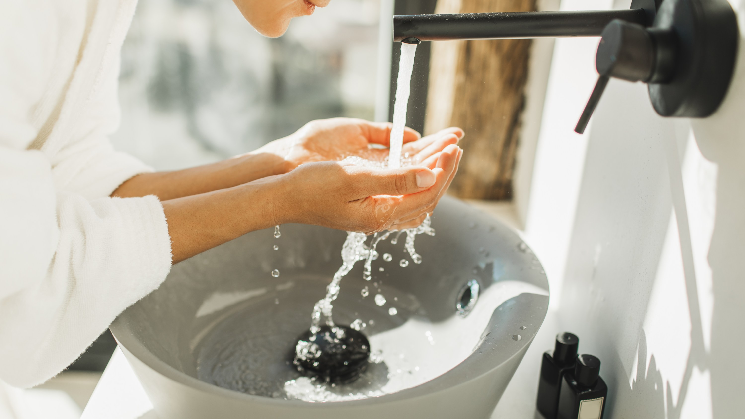 Spa treatment. Bathroom with white walls full of sun light. Black metal furniture. Grey stone sink. Person in bathrobe. Trendy interior design
