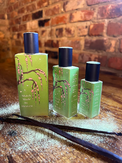 Sorcellerie Apothecary Match Made In Heaven Extrait de Parfum in green bottles with cherry blossoms printed on them. They sit on top of matcha powder and vanilla beans.