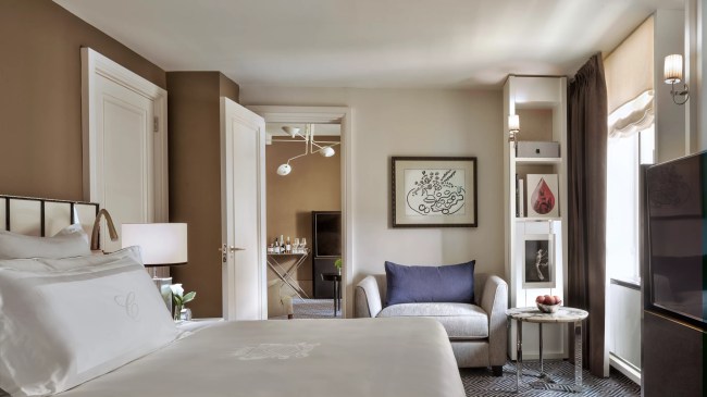 A bedroom at The Carlyle hotel, featuring a white bed, upholstered armchair, and doorway that leads to a living space with a TV