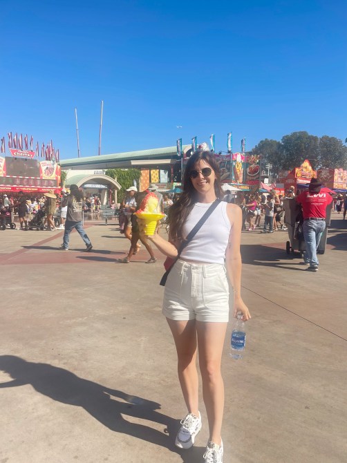 A woman wearing white shorts and a white tank top, holding a snow cone at a county fair