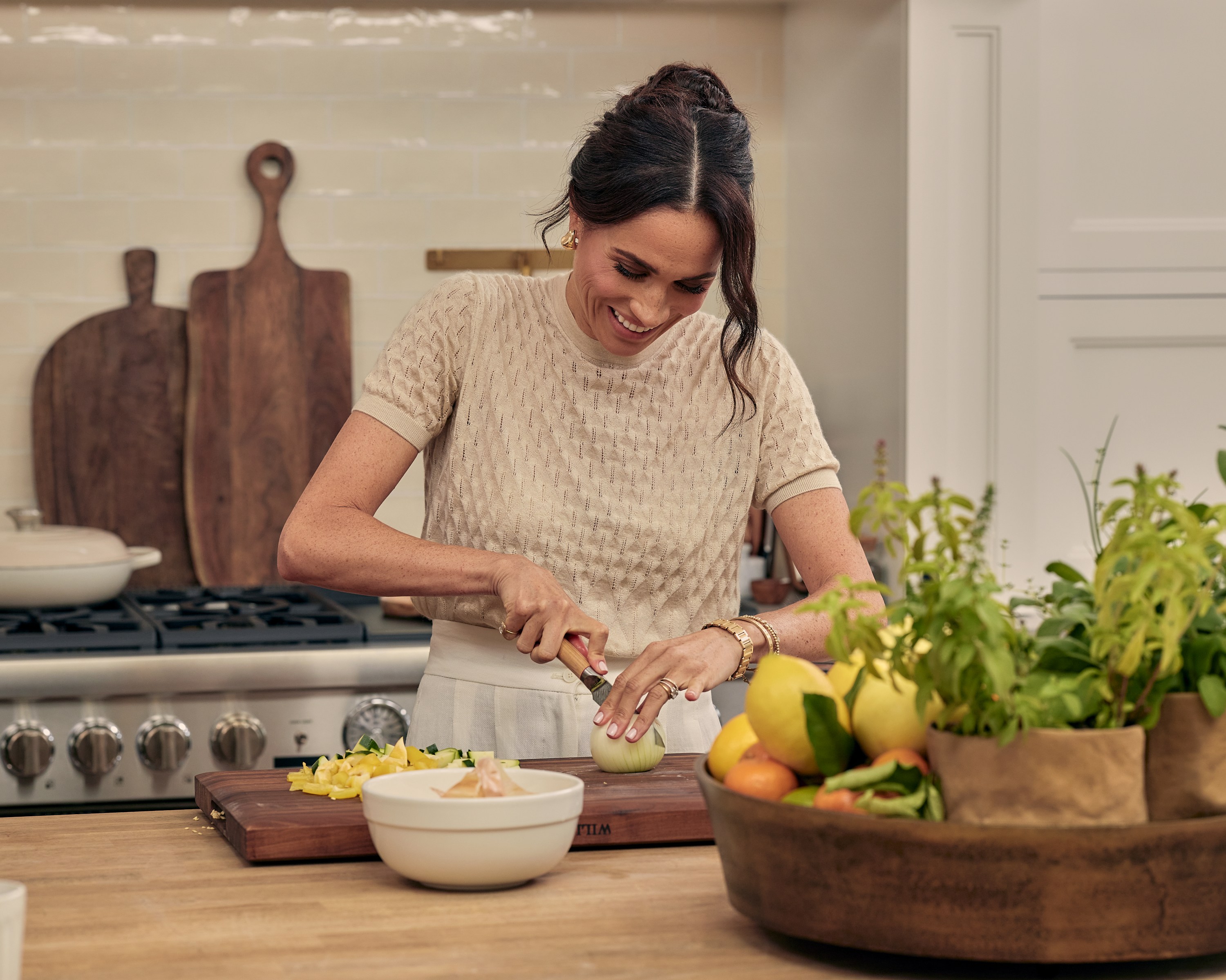 Meghan Markle in a kitchen, cutting an onion on a wooden cutting board.