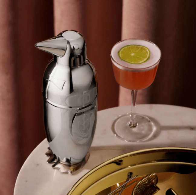 Viski Irving Penguin Cocktail Shaker in stainless steel sitting on a table next to a cocktail